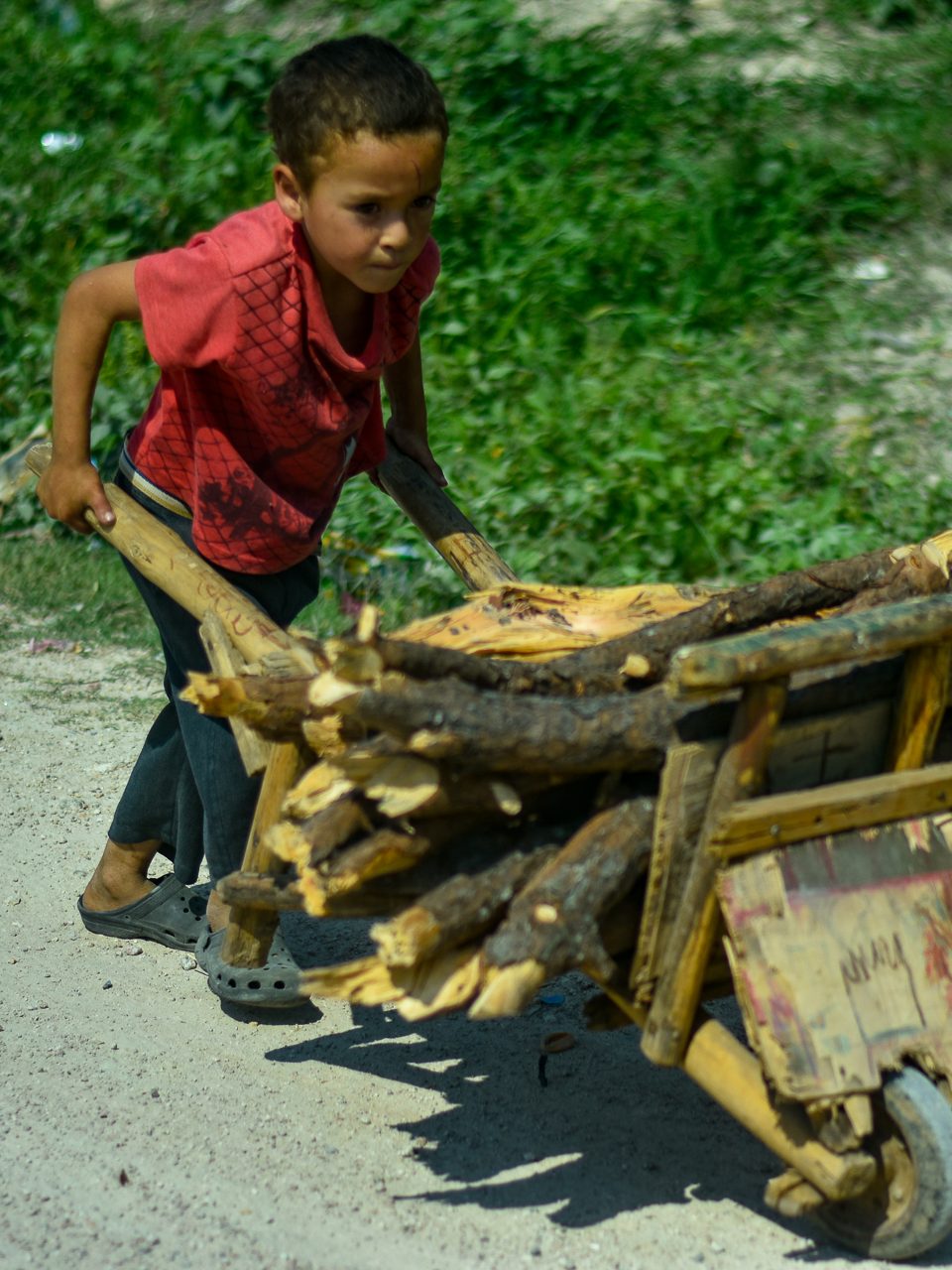 A boy pushing a wooden cart loaded with firewood.